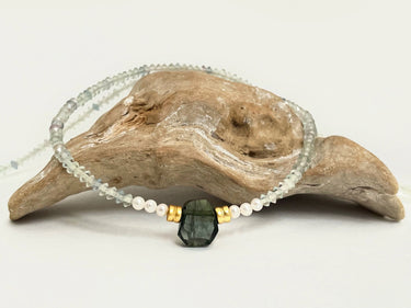 Steinkette Theresa aus kleinen , fast klaren geschliffenen Steinchen, Goldperlen und weißen Miniperlen neben einem großen dunkelgrünen Fluorit, der mittag vorne in der Kette platziert ist. Die Kette ist auf einem Holzstück platziert.